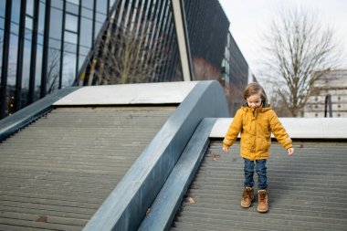 Hardal ceketli ve kot pantolonlu çocuk Vilnius 'taki bir şehir parkında modern bir ahşap binaya tırmanıyor. Kentsel kamusal alanda neşeli bir hareket ve merak anı.