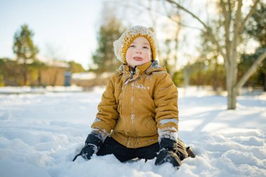 Hardal ceketli gülümseyen çocuk ve örgü şapkalı çocuk taze karda çömeliyor, soğuktan yanakları pembeleşiyor. Kış eğlencesi, açık hava ışığında basit çocukluk özgürlüğü..