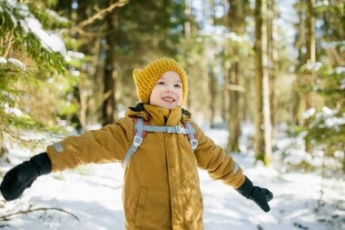 Hardal ceketli ve sarı örgü şapkalı bir çocuk sırt çantalı, güneş ışığına gülümseyen bir çocuk. Kış macera havası, vahşi doğadaki genç kaşif enerjisi..