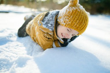 Hardal ceketli gülümseyen çocuk ve örgü şapkalı çocuk taze karda çömeliyor, soğuktan yanakları pembeleşiyor. Kış eğlencesi, açık hava ışığında basit çocukluk özgürlüğü..