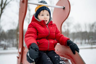 Kırmızı ceketli ve ponpon şapkalı çocuk karlı bir oyun parkından kayıyor, heyecandan sırıtıyor. Soğuk şehir havasında basit kış neşesi, hareket ve kahkaha.