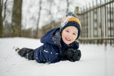 Donanma ceketli ve örgü şapkalı bir çocuk karlı bir şehir parkında duruyor, yüzü kar taneleriyle kaplanmış. Sakin bir kış anı, sakin bir varlık yumuşak şehir karları arasında.