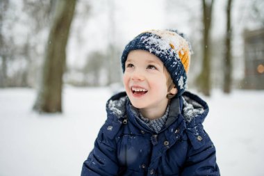 Donanma ceketli ve örgü şapkalı bir çocuk karlı bir şehir parkında duruyor, yüzü kar taneleriyle kaplanmış. Sakin bir kış anı, sakin bir varlık yumuşak şehir karları arasında.