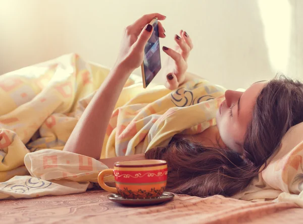 Mladá žena s chytrým telefonem ráno ležící v posteli . — Stock fotografie
