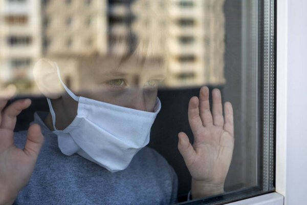 Маленький мальчик в защитной маске с тоской смотрит в окно на открытом пространстве. Способ жизни во время эпидемии.