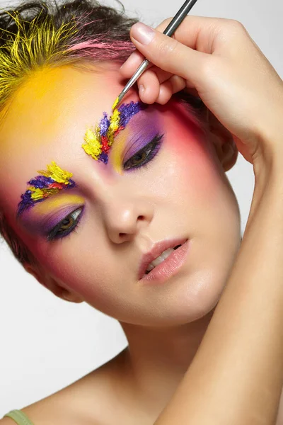 Female portrait with unusual face art makeup. Paint on brows, hair and around eyes. Artist\'s hand with paintbrush painting beautiful girl\'s brows make-up.