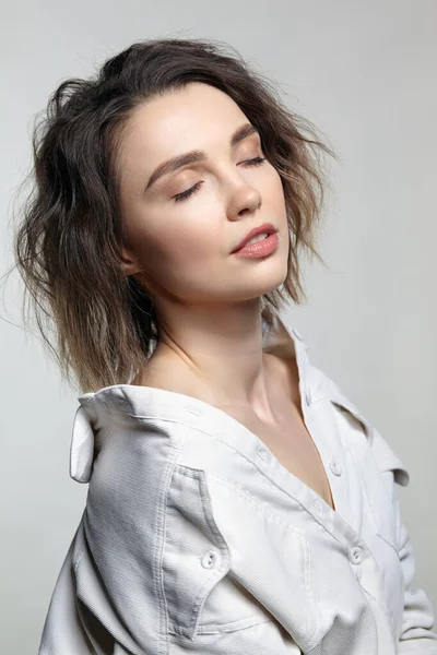 Portrait of young woman with eyes closed on gray background. Female posing in milky white micro-corduroy shirt.