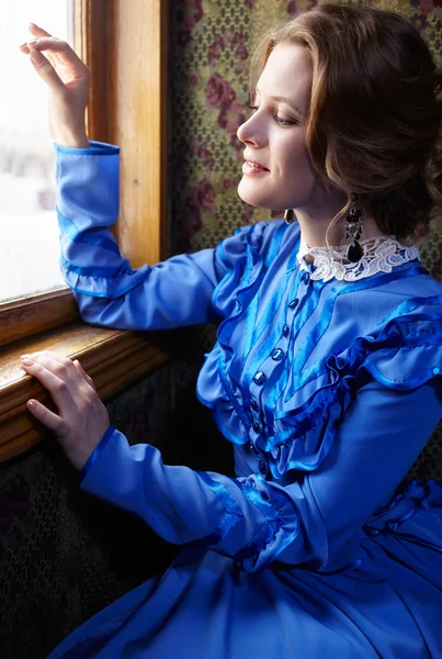 Young woman in blue vintage dress looking out the window in coup