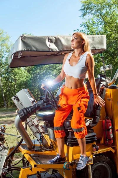 Sexy blonde Frau in orangefarbenen Overalls bedient den Traktor. — Stockfoto