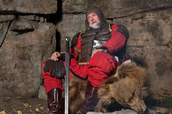 Old medieval King in armor with sword is sitting on furs