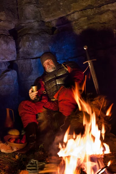 Old medieval King in armor with dish is sitting on furs near the — Stock Photo, Image