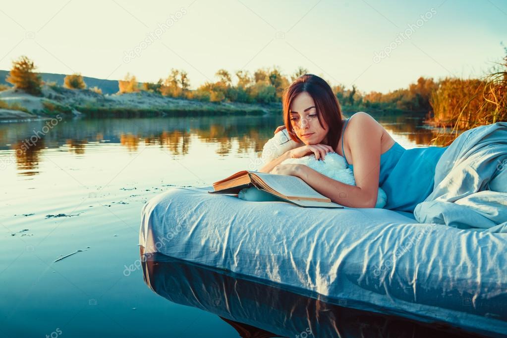 Pretty woman is laying on the floating bed and reading a book. S