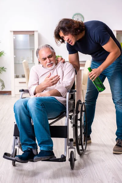 Homem velho em cadeira de rodas e jovem mau cuidador dentro de casa — Fotografia de Stock