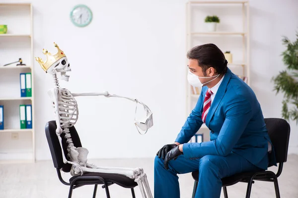 Молодой бизнесмен и скелет в офисе в пандемической концепции — стоковое фото