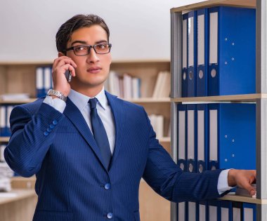 Dosyalarla rafın yanında duran genç adam.