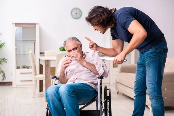 Homem velho em cadeira de rodas e jovem mau cuidador dentro de casa — Fotografia de Stock