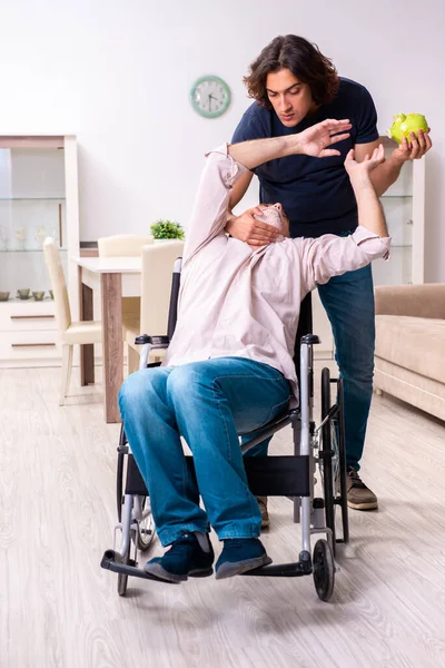 Старик в инвалидной коляске и молодой плохой сиделка в помещении — стоковое фото