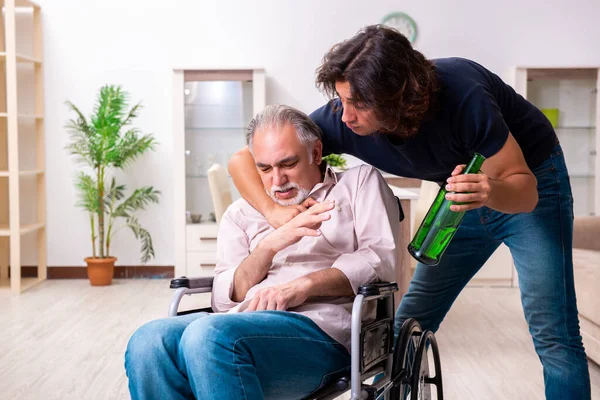 Vecchio in sedia a rotelle e giovane cattivo caregiver al chiuso — Foto Stock