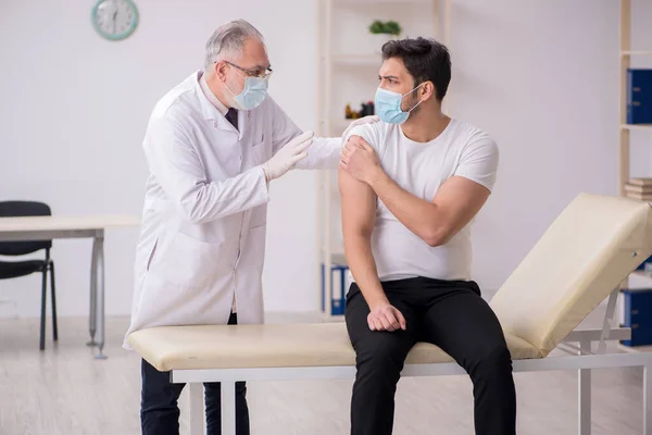 Jonge mannelijke patiënt bezoekt oude mannelijke arts in vaccinatiecentrum — Stockfoto