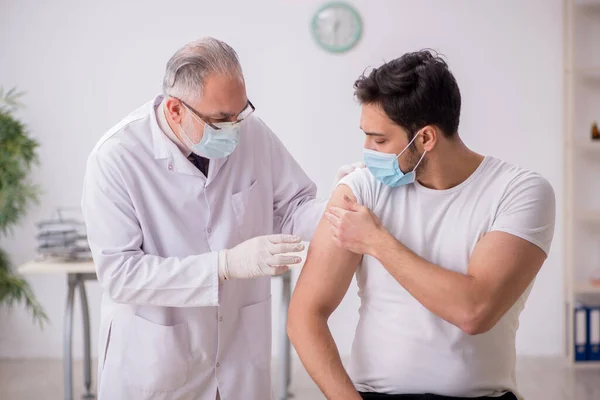 Jonge mannelijke patiënt bezoekt oude mannelijke arts in vaccinatiecentrum — Stockfoto