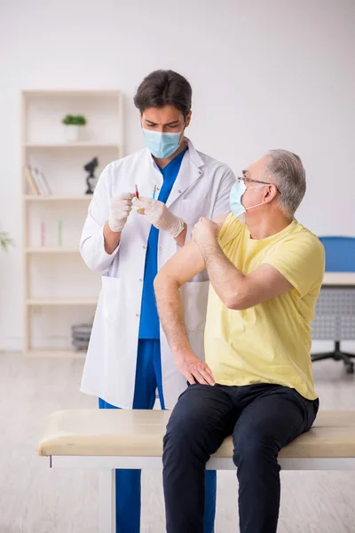 Oude mannelijke patiënt visting jonge mannelijke arts in vaccinatie concep — Stockfoto