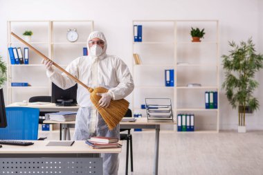 Yaşlı müteahhit elinde süpürgeyle ofisi temizliyor.