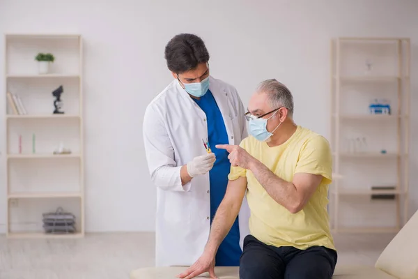 Oude mannelijke patiënt visting jonge mannelijke arts in vaccinatie concep — Stockfoto