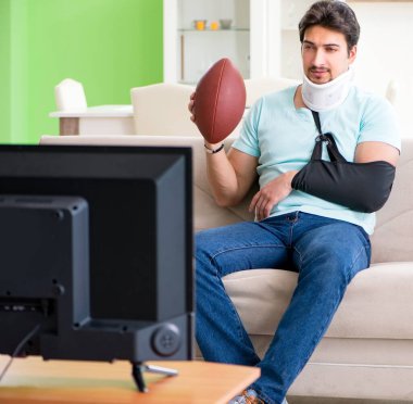 Boynu ve kolu yaralanmış bir adam televizyonda Amerikan futbolu seyrediyor.