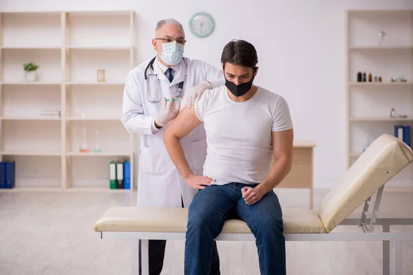 Jonge mannelijke patiënt op bezoek bij oude arts in vaccinatieconcept — Stockfoto