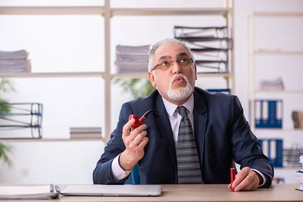 Old male employee smoking pipe at workplace — Stock Photo, Image