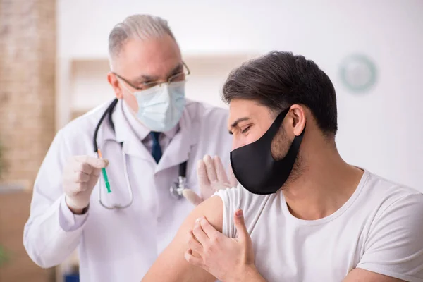 Jonge mannelijke patiënt op bezoek bij oude arts in vaccinatieconcept — Stockfoto