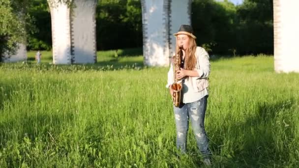 Chica en sombrero y pantalones vaqueros rasgados juega saxofón cerca de acueducto — Vídeos de Stock