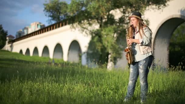 Chica en sombrero y pantalones vaqueros rasgados juega saxofón cerca de acueducto — Vídeos de Stock