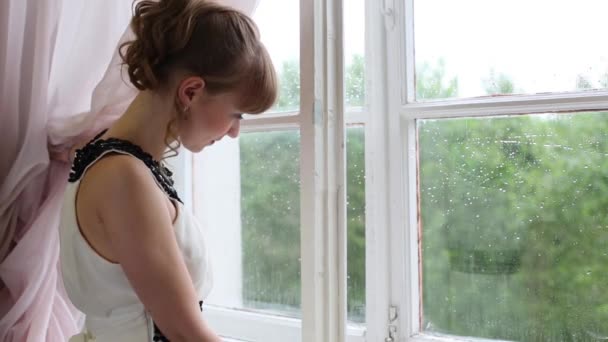 Hermosa chica triste en vestido blanco mira en la ventana durante la lluvia — Vídeo de stock