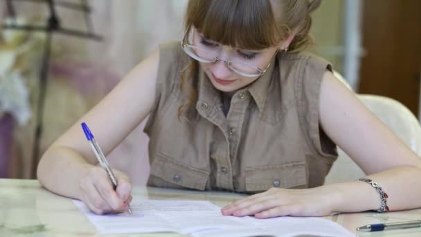 Chica bonita en gafas se sienta a la mesa y escribe en papel en la habitación — Vídeos de Stock