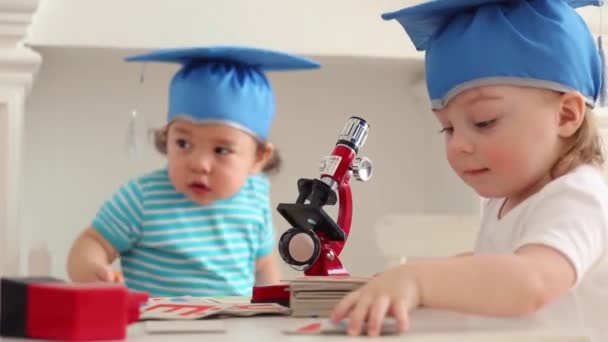 Bebés en sombreros de graduación azul juegan con microscopio en la mesa — Vídeo de stock