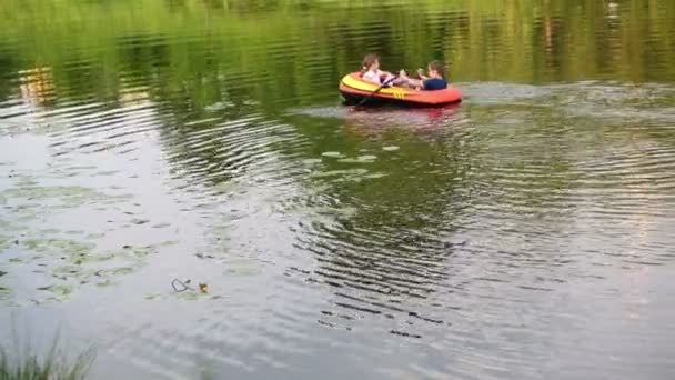 Niño y niña flotan en bote inflable con remos a lo largo del estanque . — Vídeos de Stock