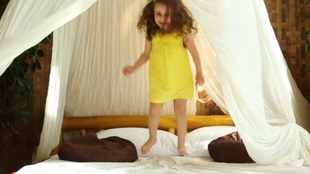 Pequeña chica rizada en vestido amarillo está saltando sobre la cama . — Vídeo de stock