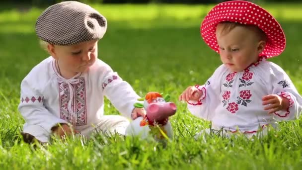 Deux bébés mignons en vêtements folkloriques jouent avec jouet vache drôle sur l'herbe — Video