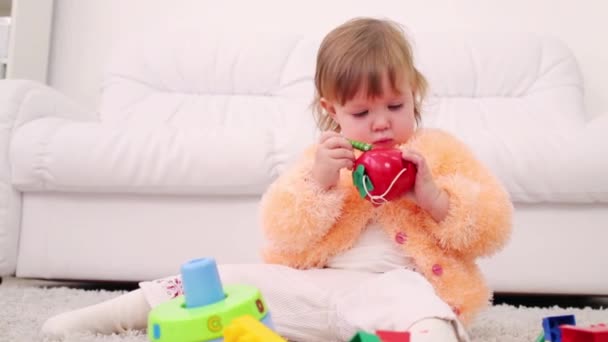 Досить маленька дівчинка сидить на килимі і тримає іграшкове яблуко з хробаком — стокове відео