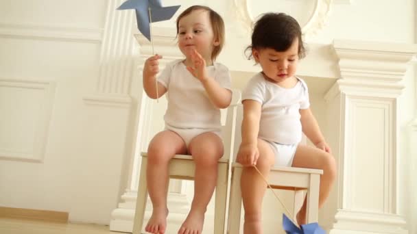 Schattige baby jongen en meisje met speelgoed windmolens zitten op houten stoelen — Stockvideo