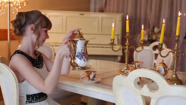 Meisje zit aan tafel met gerechten en verlichte kaarsen en giet thee — Stockvideo