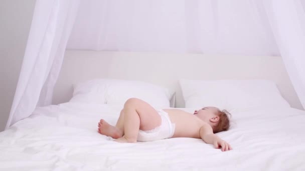 Счастливый маленький ребенок в пеленках лежит на белой кровати и поворачивается — стоковое видео