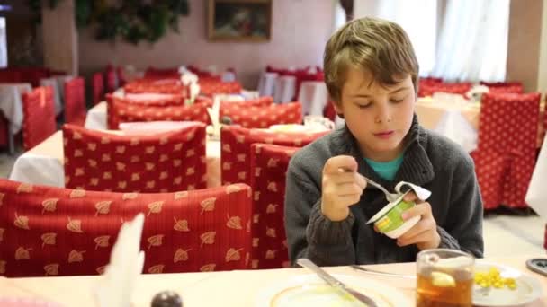 Handsome boy eats yogurt in empty canteen with red chairs — Stock Video