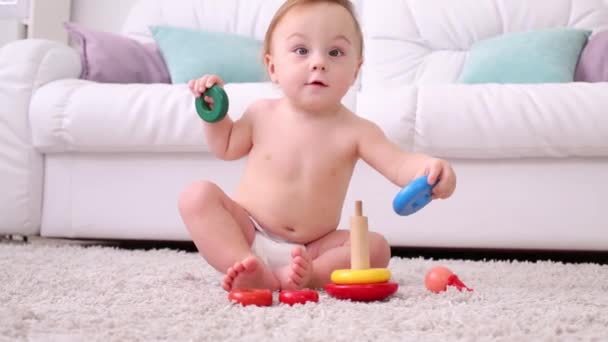 Mignon petit enfant en couches est assis sur le tapis et joue avec la pyramide — Video