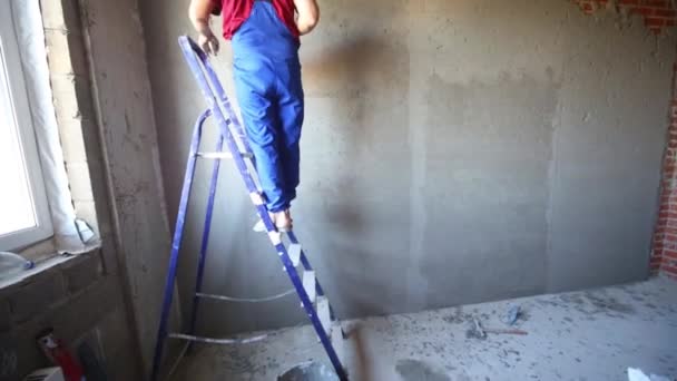 El hombre trabajador se para en la escalera y hace yeso en la pared — Vídeo de stock