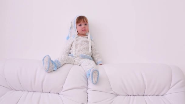 Мила дитина в капелюсі і в'язаний костюм сидить на білому дивані вдома — стокове відео