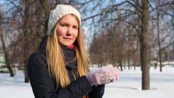 Young woman in hat holds in hands snow and blows on it in park — 비디오