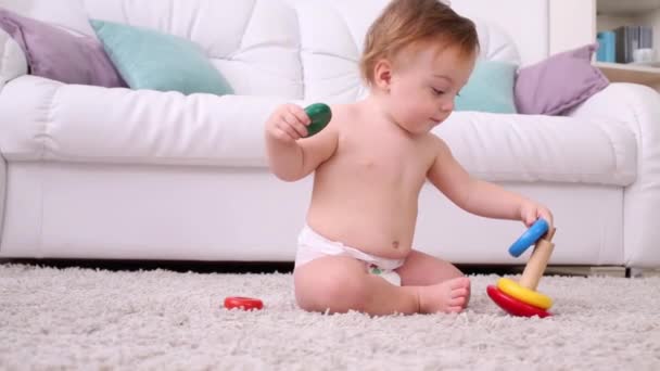 Pequeño niño en pañal se sienta en la alfombra y juega con la pirámide — Vídeo de stock