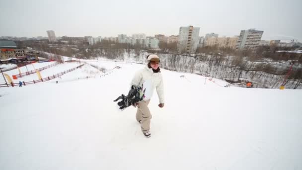 Snowboarder menina sobe a colina segurando um snowboard na mão — Vídeo de Stock